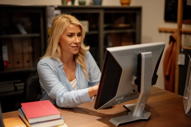 A woman sits at a desk in an office environment. She is using a computer and appears focused on her work. There are books and items on the desk around her. Administration careers jobs: the search terms to use