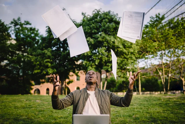 Overjoyed African American graduate tossing copies of resumes in air after learning news about successfully getting job while sitting in green park with laptop. Best resumes and cover letters courses: a buyer's guide